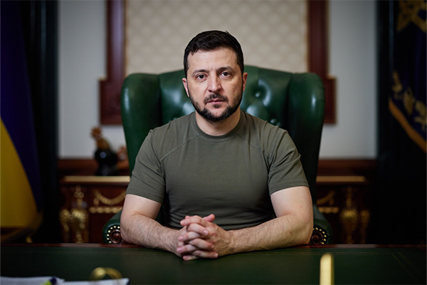President Volodymyr Zelenskyy seated in a green high-back chair, leaning forward on his desk with his hands together looking stern