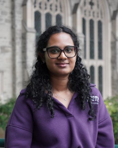 A female student with black hair and a purple sweater stands smiling in front of a wall