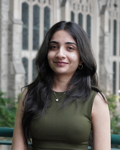 A female student in a green shirt with black hair stands smiling in an atrium with sandstone walls