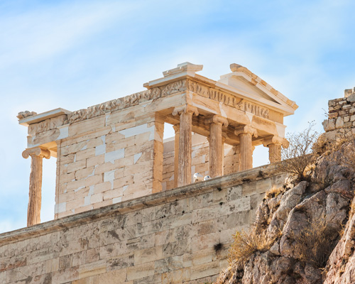 photo of a wall in Greece on the bottom. the top half of the photo shows an Ancient Greek structure with columns in the front and the back