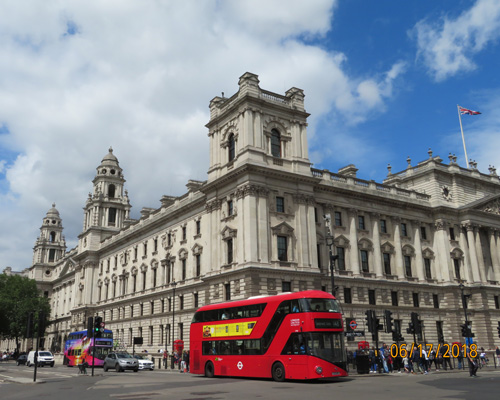 corner view of a building in the UK. Bright red double decker bus with yellow strip is in the center of the photo.