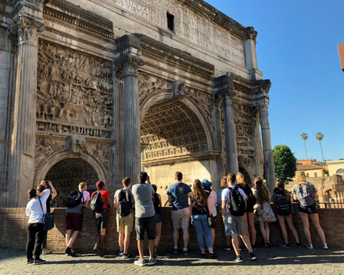 Students standing in front of an arch in Italy, backs to the camera.