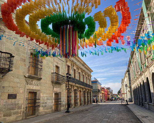Empty street in Oaxaca, Mexico. Suspended over street is a bright flag sculpture art with the colours of the rainbow. In the center, streamers hang down.