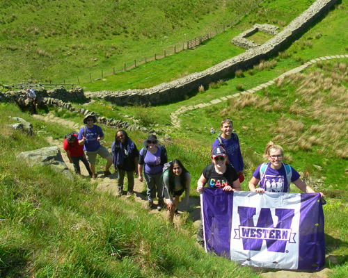 students trekking the countryside in a single file line. the two students at the front of the line are holding a purple and white Western flag.